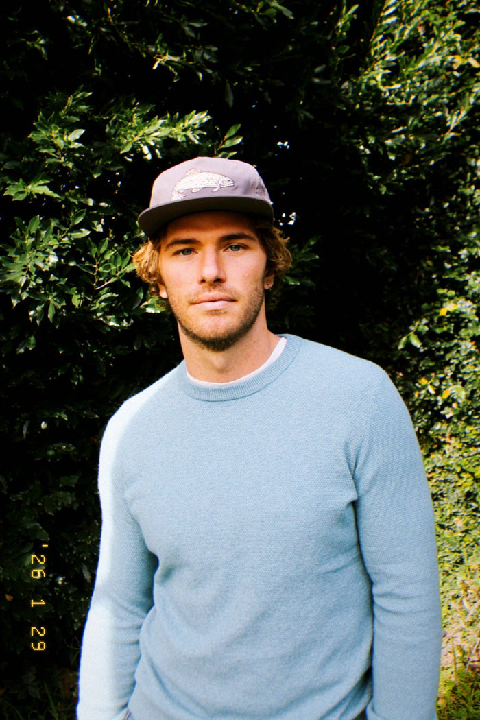 Man wearing The Trout Hat gray cap with fish logo outdoors among green foliage.