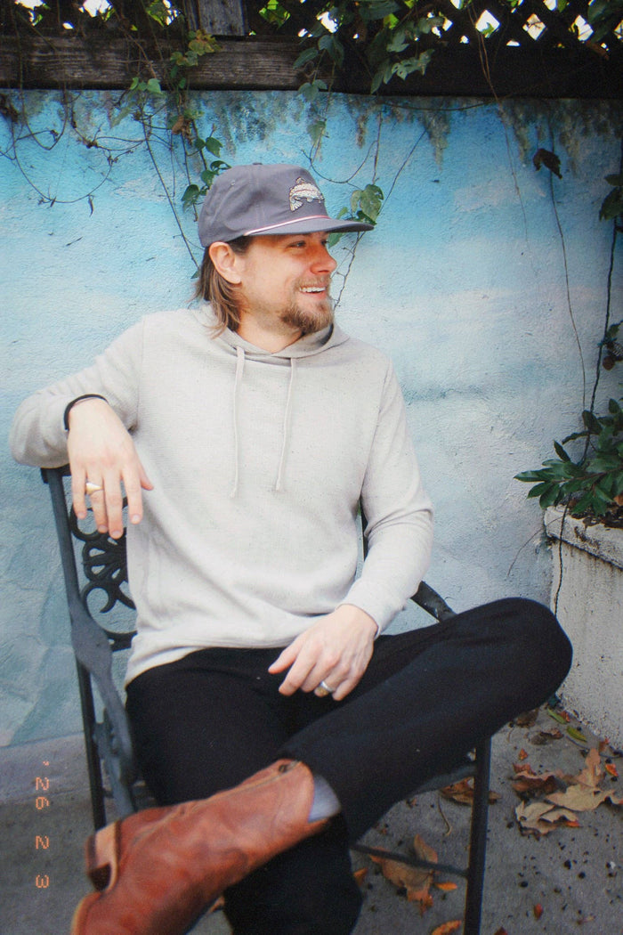 Man seated wearing The Trout Hat gray cap with fish logo and light gray hoodie against a blue wall.