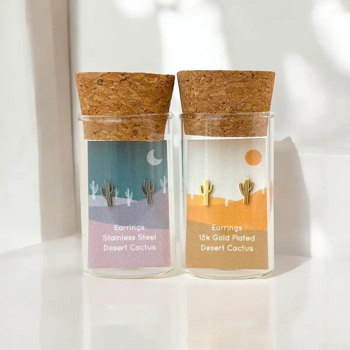 Two glass jars with cork lids contain tiny desert cactus earrings in stainless steel and 18k gold plated finishes.