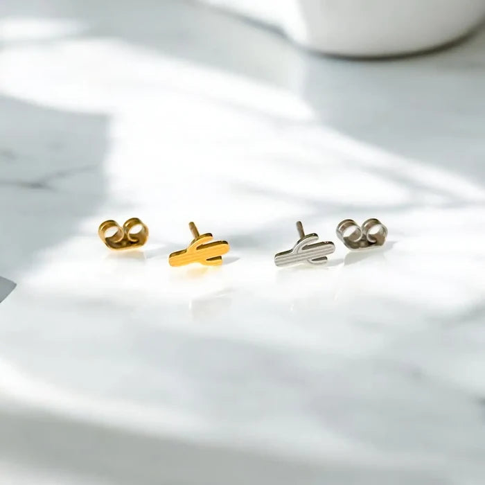 Gold and silver desert cactus stud earrings arranged on a white glossy surface with soft shadows.