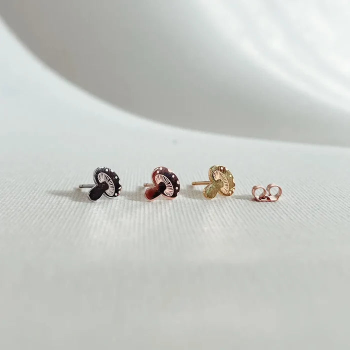 Three mushroom shaped stud earrings in black, rose gold and gold finishes with white dotted caps on a light surface