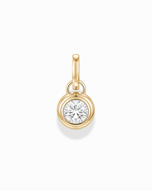 The Tove Sapphire Bezel Charm by Mod + Jo is a small 14k gold pendant featuring a round, clear lab-grown sapphire in a simple bezel setting with a gold loop, shown on a white background.