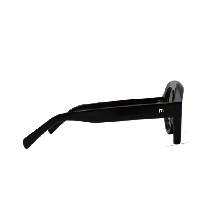 Side view of the Val Sunglasses by Elisa Johnson: black, thick-framed acetate aviator style with straight arms and a small gold M logo on the temple. UV400 protection. Minimalistic white background. Brand: Elisa Johnson.