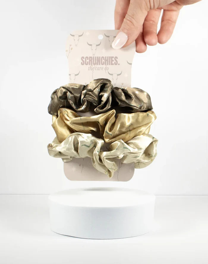 Three metallic scrunchies in bronze and green tones on a pale card with Scrunchies branding