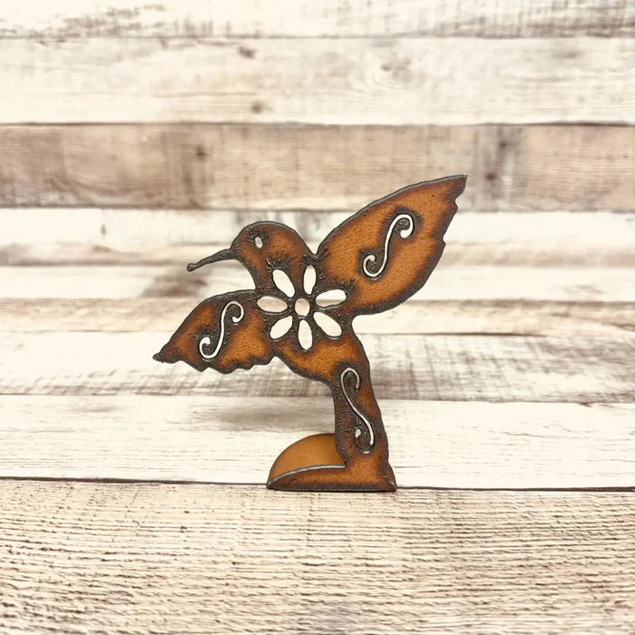 Rusty orange metal hummingbird cutout standee with a central flower and white scroll accents on a wooden surface.