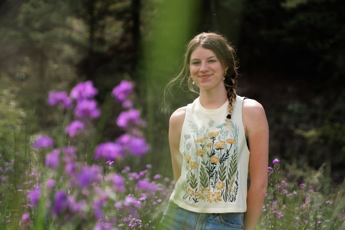 Model wearing Wild Field Ladies Crop Tank in a sunny meadow with purple flowers.
