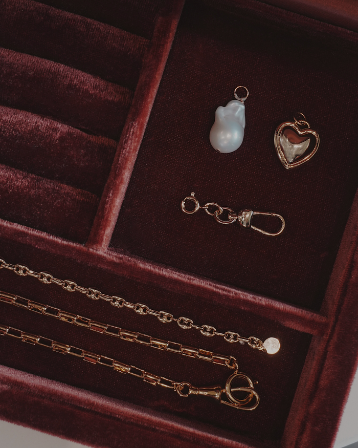 Jewelry laying in a jewelry box including a three gold chain, a heart pendant, a pearl charm. and a charm clip, all laying on a dark red velvet surface.