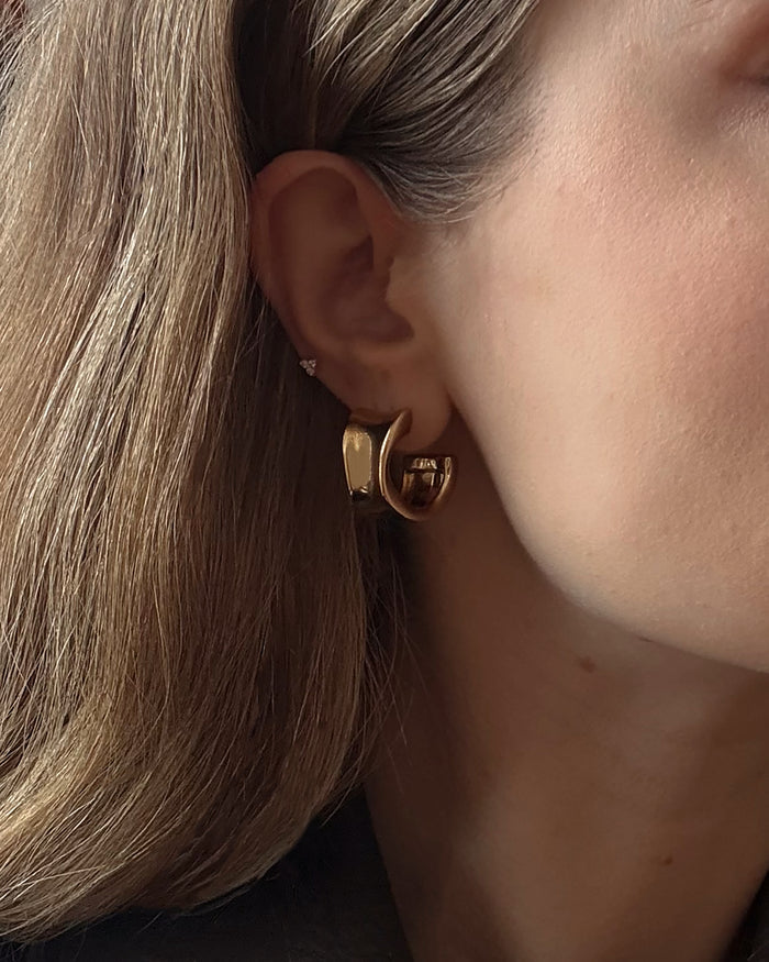 Close-up of a person's ear wearing Zara Volume Hoops and a small stud earring. The person has light brown hair, fair skin, and the background is softly blurred.